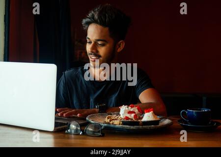 Bart asiatischen Mann in Freizeitkleidung arbeiten mit Computer und essen Kuchen mit Tee in Cafe.Student männlich Chat sozialen Netzwerk Hausaufgaben zu tun Stockfoto