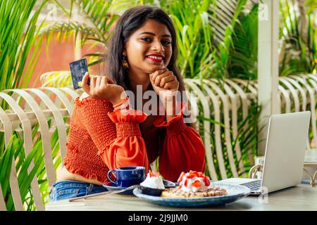 Weibliche Kundin, die für Kaffee und Pfannkuchen mit Kreditkarte bezahlt, die mit einem Laptop sitzt und am freitag online einkaufen geht Stockfoto