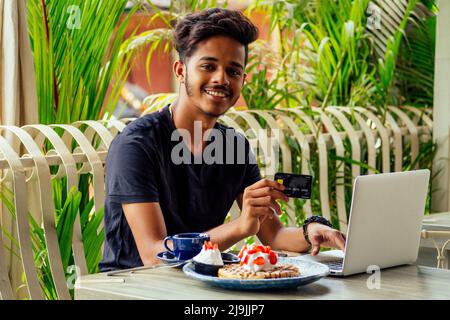 Männlicher Kunde, der für Kaffee und Pfannkuchen mit Kreditkarte bezahlt, der mit einem Laptop sitzt und am schwarzen freitag online einkaufen geht Stockfoto