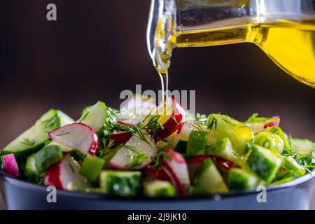 Gießen von Olivenöl auf gesunde frische Gemüsesalat, Nahaufnahme. Köstlicher Salat aus Gurken, Rettich, grünen Zwiebeln, Dill und Olivenöl. Co Stockfoto
