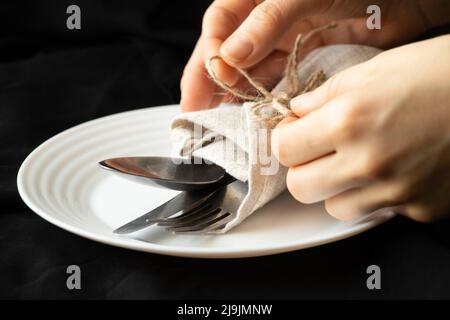 Vintage alte Gabel und Messer in Servietten und Händen binden eine Seilschleife auf einer Serviette, liegen auf einem Teller auf einer schwarzen Tischdecke auf dem Tisch, Wortdienen, Silberkrieg Stockfoto