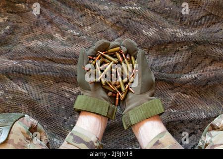 Ukrainischer Soldat in Handschuhen hält Kugeln aus einem Maschinengewehr auf einem Tarnhintergrund, Waffen der ukrainischen Armee, Krieg in der Ukraine 2022 Stockfoto