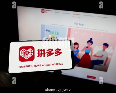 Person, die Mobiltelefon mit dem Logo des chinesischen E-Commerce-Unternehmens Pinduoduo Inc. Auf dem Bildschirm vor der Business-Webseite hält. Konzentrieren Sie sich auf die Telefonanzeige. Stockfoto