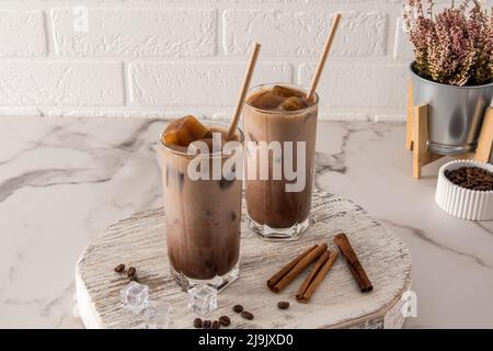 Zwei Gläser mit kaltem Latte-Kaffee oder mit einem Schokoladengetränk und Eiswürfeln, Zimt auf einem weißen Holzbrett und einem Marmortisch Stockfoto