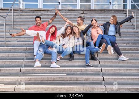 Eine Gruppe fröhlicher, multiethnischer junger Männer und Frauen in lässigen Outfits, die fröhlich lächeln und auf die Kamera schauen, während sie auf der Treppe sitzen und sich nach ihrem erfolgreichen Studium umarmen. Stockfoto