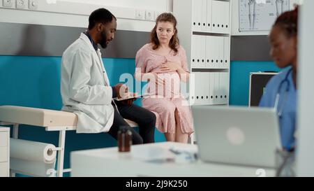 Gesundheitsspezialist, der eine Kontrollkonsultation mit einem erwartenden Patienten im Büro macht. Männlicher Arzt mit digitalen Tablette, um Notizen zu machen, die medizinische Beratung für Frau mit Schwangerschaft Bauch. Stockfoto