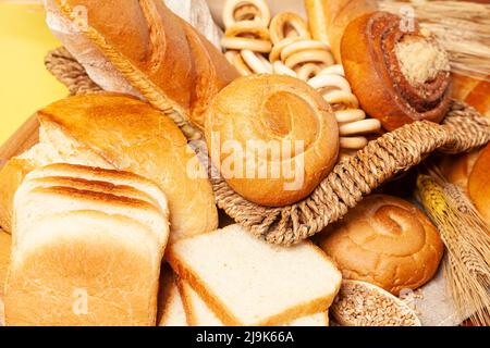 Kulinarischer Hintergrund mit Brot, Brötchen, trocknenden Bagels, Weizenohren in einem Korbkorb, auf Gelb Stockfoto