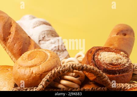 Kulinarischer Hintergrund mit Brot, Brötchen, trocknenden Bagels, Weizenohren in einem Korbkorb, auf Gelb Stockfoto