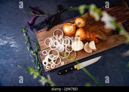 Eine Menge Zwiebeln in einem Korb und Teller auf einem dunklen und eleganten dunklen Strukturhintergrund. Zwiebeln und Scheiben auf Holzschneidebrett anbraten. Stockfoto