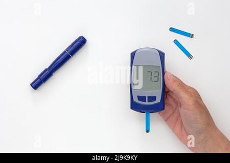 Draufsicht auf das von einer Frau in der Hand geschnittene Glucometer mit 7,3 Ergebnissen auf dem Display mit blutigem Teststreifen im Inneren, Lanzette und Teststreifen auf weißem Hintergrund Stockfoto