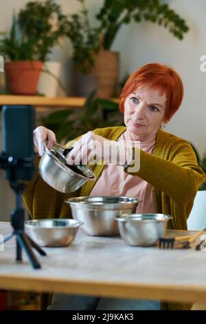 Reife Frau, die ihr Mobiltelefon zum Streamen benutzt, sitzt am Tisch und erzählt vom Pflanzen Stockfoto