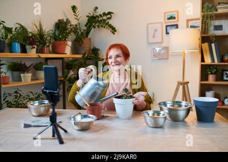 Reife Frau wässern Topfblume mit Gießkanne und erzählen über Pflanzen, Anhänger während Streaming auf Handy Stockfoto