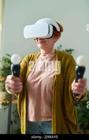 Ältere Frau in legerer Kleidung, die drahtlose Brillen und Joysticks verwendet, um zu Hause virtuelles Spiel zu spielen Stockfoto