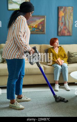 Afrikanisches Mädchen, das den Teppich im Wohnzimmer mit einem kabellosen Staubsauger säubert, um älteren Frauen bei der Hausarbeit zu helfen Stockfoto