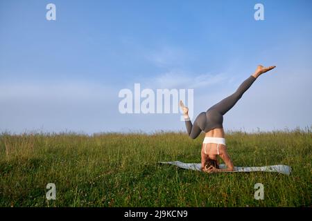 Starke junge Frau im Sportoutfit üben Kopfstand Position zwischen Sommer Berge. Aktive weibliche Person, die während der Morgenstunden im Freien Akro-Yoga macht. Stockfoto