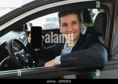 Hübscher junger Autoverkäufer, der im Autosalon sitzt und leeres Mobiltelefon mit Modell für Design im Autohaus zeigt Stockfoto
