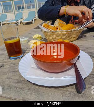 Köstliche cremige italienische Tomatensuppe mit Pomodori, serviert mit einem Korb mit Baguette und Kräuterbutter und einem Glas Eistee ohne Marke daneben Stockfoto