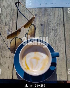 Köstlicher und farbenfroher cremiger Cappuccino, der in einer blauen Tasse auf einem Terrassentisch mit Sonnenbrille und einem Menü daneben serviert wird Stockfoto