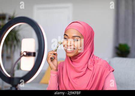 Fröhliche schwarze Dame macht Gesichtsmassage mit Steinwalze vor dem Handy, macht Video für Beauty-Blog Stockfoto