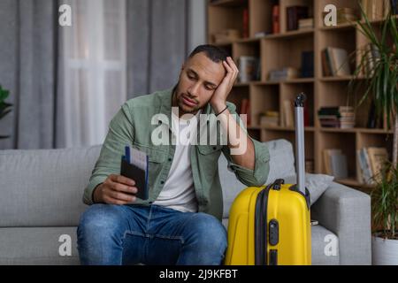 Verärgert afroamerikanischen Mann mit Pass und Flugtickets auf dem Sofa sitzen neben gepackten Koffer, verpasste seinen Urlaub Stockfoto