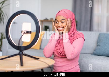 Glückliche schwarze muslimische Bloggerin, die von zu Hause aus sendet, die Handykamera anschaut, lächelt und die Wangen berührt Stockfoto