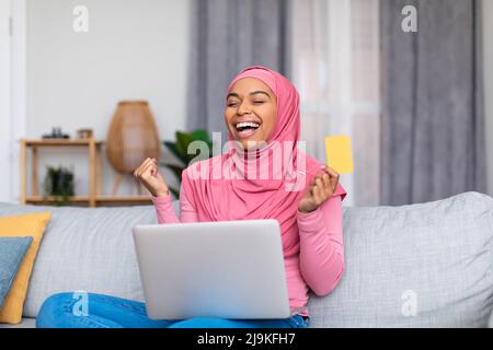 Überglücklich schwarze Frau in Hijab Einkaufen im Online-Shop auf Laptop, halten Kreditkarte und gestikulieren JA Stockfoto
