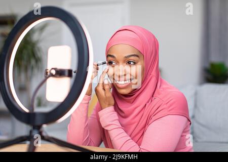 Online-Make-up-Tutorial. Glückliche schwarze Frau, die Video für ihren Internet-Blog macht und vor dem Smartphone sitzt Stockfoto