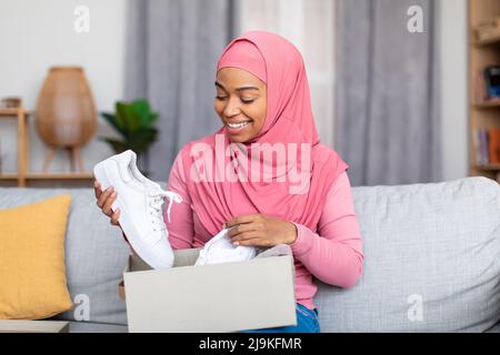 Muslimische Kundin glücklich mit erhaltenen Sneakerschuhen, dem Öffnen der Box aus dem Online-Shop und dem Betrachen des gelieferten Artikels Stockfoto