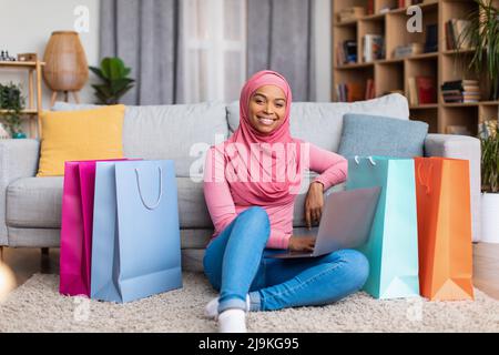 Positive schwarze muslimische Dame mit Laptop und Einkaufstaschen, die zu Hause über das Internet einkaufen, auf dem Boden sitzen und lächeln Stockfoto