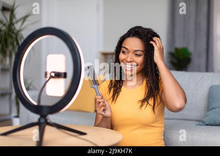 Haarpflege und Blogging Konzept. Glückliche afroamerikanische Frau, die Kritik auf dem Haarkamm aufzeichnete, indem sie Mobiltelefon auf der Ringlampe verwendete Stockfoto