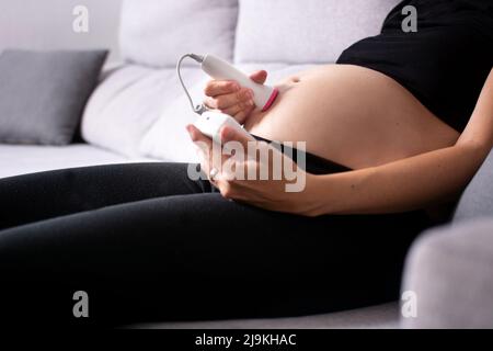 Eine Schwangerin mit einem fetalen doppler hört dem Baby zu Hause auf dem Sofa zu Stockfoto