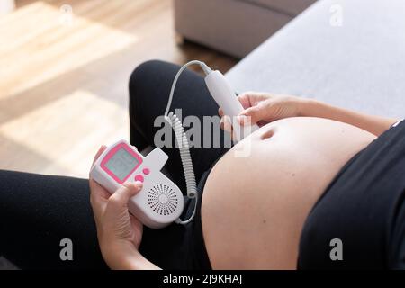 Eine Schwangerin mit einem fetalen doppler hört dem Baby zu Hause auf dem Sofa zu Stockfoto