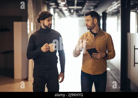 Männliche Kollegen diskutieren über das Geschäftsprojekt und nutzen einen Tablet-pc, während sie während der Kaffeepause im Büro in der Firmenhalle spazieren gehen Stockfoto