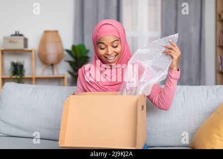 Konzept für schnelle Lieferung. Glückliche muslimische Frau erhielt Paket, Auspacken Karton und lächelnd, sitzen auf dem Sofa zu Hause Stockfoto