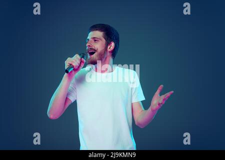 Hübscher Mann im weißen T-Shirt, der am Mikrofon singt, isoliert auf dunkelblauem Studiohintergrund, Konzept menschlicher Emotionen, Gesichtsausdruck, Umsatz, Anzeige Stockfoto