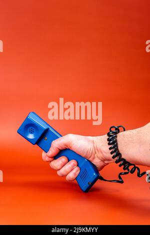 Hand, die ein blaues Telefon mit dem Telefonkabel um den Arm auf einem orangefarbenen Hintergrund hält. Stockfoto