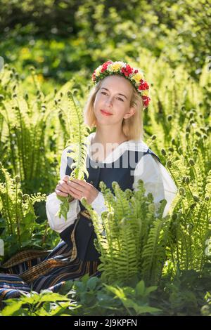 Junge Frau in Nationalkleidung und Kranz vor dem Hintergrund eines grünen Farns. LIGO Lettischer Feiertag Stockfoto