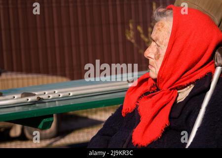 Defokussieren sie die ältere ukrainische Frau in einem roten Schal mit Krücken, die draußen sitzen. Frau 90 Jahre alt. Einzelperson. Nahaufnahme Porträt. Krank und heimatverbunden. Aus Stockfoto