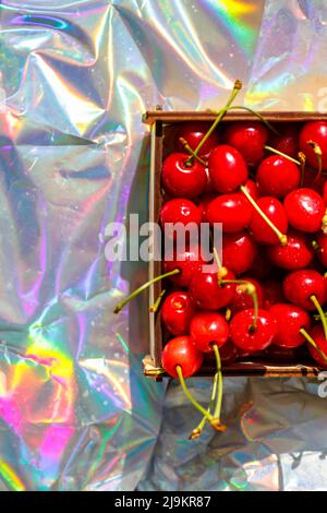 Unschärfe-Nahaufnahme-Box, Crape von dunkelroten, nassen Süßkirschen mit Schwanz und Blättern auf modernem Hologramm-Hintergrund. Früchte und Beeren. Ernte und Stockfoto