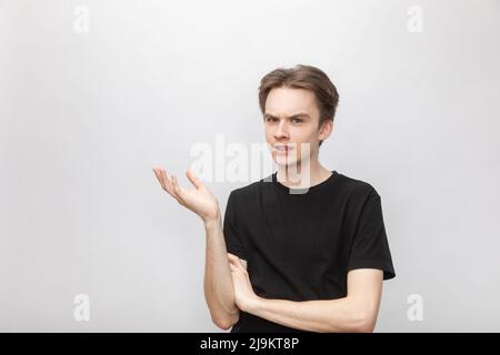 Porträt eines zweifelhaften jungen Mannes, der in einem schwarzen T-Shirt die Hand hält, während er eine fragende Geste mit arglistigen Blick auf die Kamera macht. Studio auf grauem Hintergrund aufgenommen Stockfoto