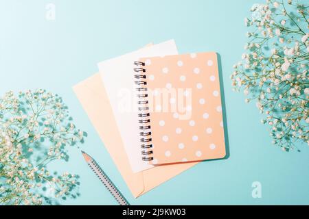 Notizblock und Umschlag mit leerem Blatt, Bleistift auf blauem Hintergrund mit Gypsophila-Blüten im Sonnenlicht. Draufsicht, flach liegend. Stockfoto