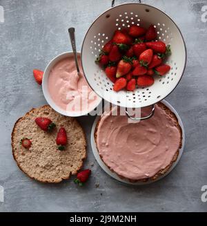 Erdbeerkuchen in Bearbeitung. Nahaufnahme von Erdbeerherb-Zutaten auf einem Tisch. Frische Beeren, Milchcreme, Kuchenlagen Draufsicht. Stockfoto
