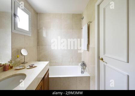 Toilette mit Duschkabine, cremefarbenen Marmorfliesen und Arbeitsplatte mit weißem Waschbecken auf Holzschrank mit Gittern Stockfoto