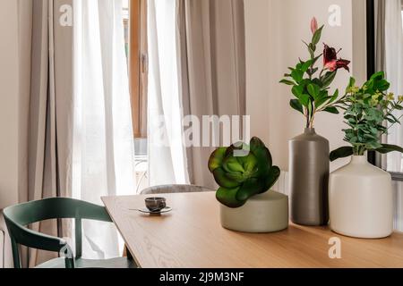 Porzellanvasen mit dekorativen Pflanzen und einer Tasse Kaffee auf einem rechteckigen Holztisch und einem Fenster mit Vorhängen und Netzvorhängen Stockfoto