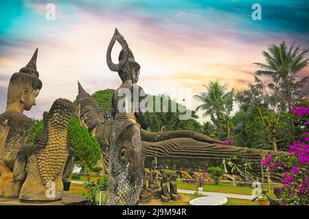 Viele buddhistische und hinduistische Steinskulpuren, Palmen, stimmungsvoller Sonnenuntergang, lila Blumen - Buddha Park (Wat Xieng Khuan), Vientiane, Laos Stockfoto