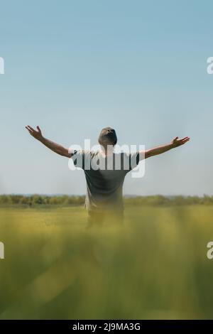 Zufriedener erfolgreicher Landwirt, der am sonnigen Frühlingstag in siegreicher Pose im unreifen Gerstenanbaugebiet die Hände hebt. Rückansicht eines grün getragens Farmarbeiters Stockfoto