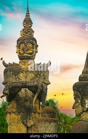 Furchteinflößende mythologische hinduistische mystische Kreatur Steinskulptur und buddha Kopf, dramatischer Sonnenuntergang Himmel, Palmen - Buddha Park (Wat Xieng Khuan), Vientiane, L Stockfoto