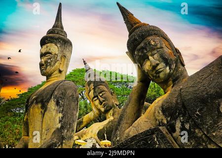 Drei mystische Buddha-Kopfstatuen aus Stein, dramatischer Sonnenuntergang am Himmel - Buddha Park (Wat Xieng Khuan), Vientiane, Laos Stockfoto