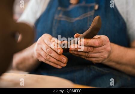 Beschnittene, nicht erkennbare weibliche Keramikerin in Schürze, die in der Werkstatt ein Stück Ton in der Hand formt Stockfoto