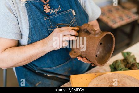 Geknöpfte, nicht erkennbare weibliche Keramikerin in Schürze, die in der Werkstatt ein Stück Tontopf in der Hand formt Stockfoto